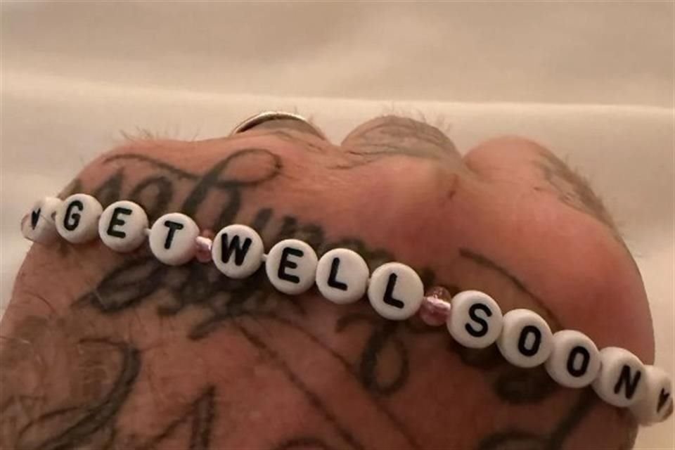 David aparece descansando en una cama blanca mientras sostiene una pulsera de amistad con la frase 'mejórate pronto'.