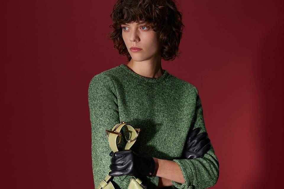 Un llamativo suéter en color verde claro es usado por una joven de mediana estatura, utilizando solo como accesorios sus guantes y falda negra junto a un bolso en tono pistache.